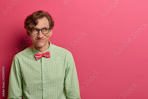 Photo of funny joyful man wears elegant green shirt and bowtie, transparent glasses, has cheerful positive look aside, plans something in mind, isolated on pink background, copy space for text