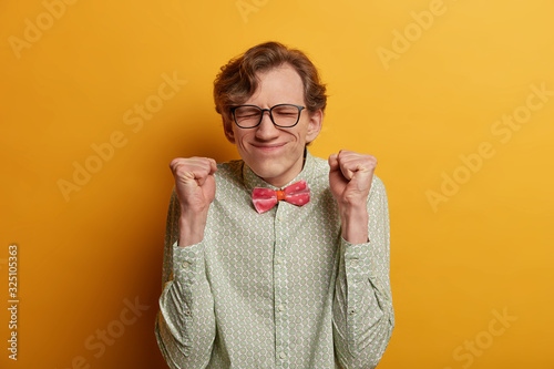 Ecstatic male nerd raises clenched fists, believes in success, closes eyes and awaits announcement of results, participates in contetst, hopes to win, expresses impatience, dressed formally. photo