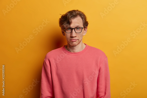 Bored unimpressed male student looks seriously at camera, has fatigue expression, wears optical glasses and pink jumper, sighs from tiredness, isolated on yellow background. Face expressions © wayhome.studio 