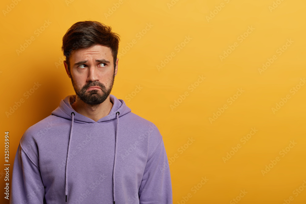 Indoor shoto of gloomy dissatisfied man feels depressed and lonely, being in despair, thinks over some problems, frowns face, dressed casually, poses against yellow background, free space aside
