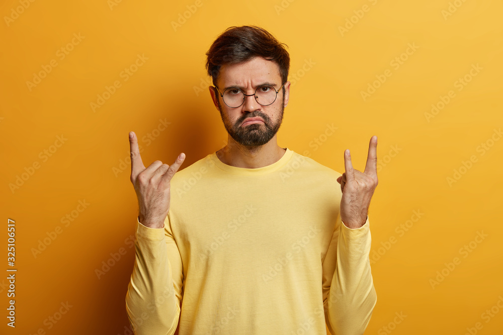 Serious looking bearded man shows cool rock n roll gesture, makes heavy metal sign, being real rocker, wears optical glasses and jumper poses against yellow background attends concert of favorite band Serious looking bearded man shows cool rock n roll gesture, makes heavy metal sign, being real rocker, wears optical glasses and jumper poses against yellow background attends concert of favorite band