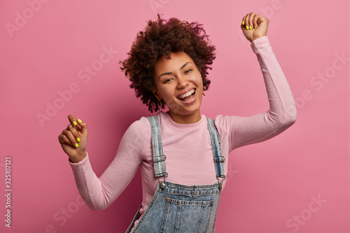 Fototapeta Naklejka Na Ścianę i Meble -  Upbeat dark skinned African American female model dances and feels joyful, wears turtleneck and denim sarafan, smiles broadly, has fun and moves with rhythm of music, isolated over pink background