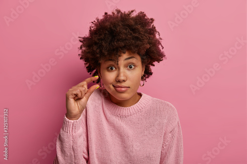 Headshot of curly haired woman discusses something very small, shapes something very tiny, purses lips, dressed in casual jumper, isolated on pink background, asks tiny object, makes small gesture photo