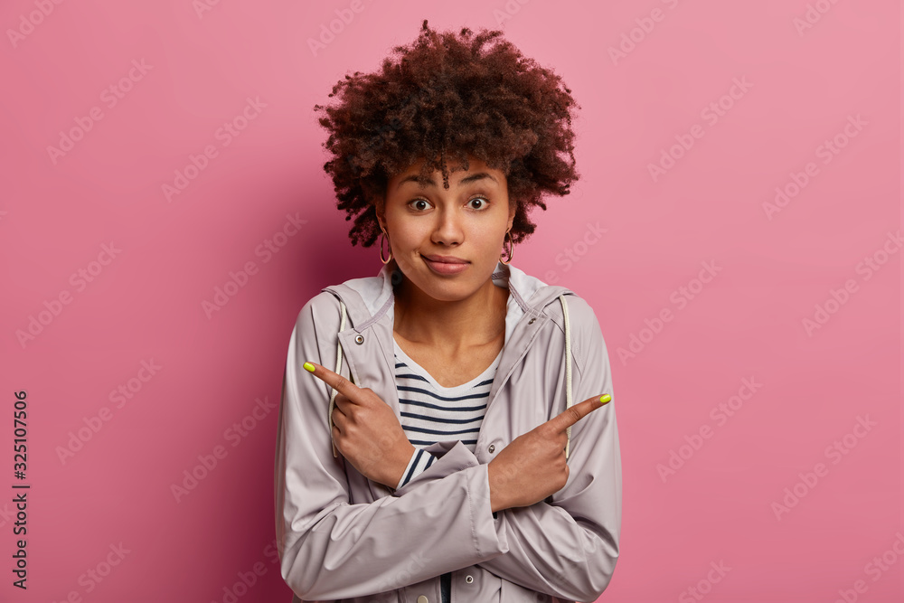 Embarrassed Afro American woman has to make difficult choice, points left and right, looks with questioned hesitant expression, shows two ways, wears grey anorak, isolated on pink background Embarrassed Afro American woman has to make difficult choice, points left and right, looks with questioned hesitant expression, shows two ways, wears grey anorak, isolated on pink background