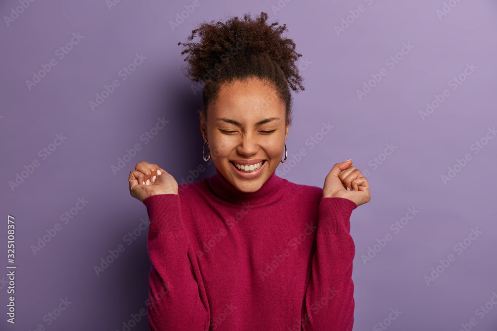 Cheerful glad ethnic girl cheers something with clenched fists, smiles broadly, gestures actively, happy with good luck or promotion at work, dressed casually, isolated over purple background. Cheerful glad ethnic girl cheers something with clenched fists, smiles broadly, gestures actively, happy with good luck or promotion at work, dressed casually, isolated over purple background.