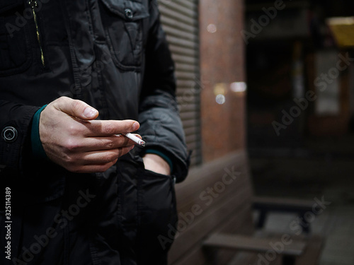 Male hand holds a cigarette. Concept of smoking addiction, cessation. Young man in black jacket with cigarette on the street.