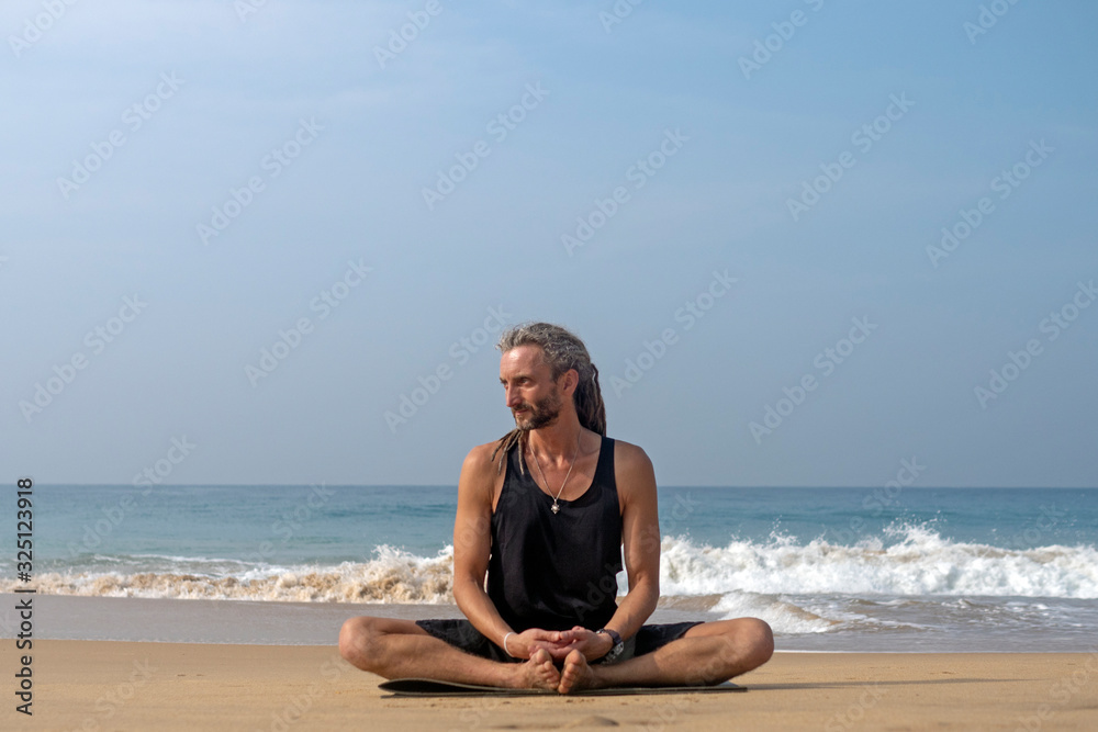 A man with dreadlocks on his head does yoga on the beach overlooking the ocean. Sri Lanka.