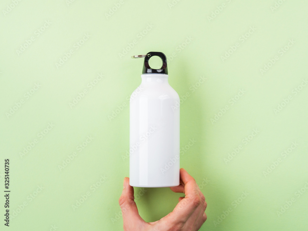 Zero waste concept - white metal reusable water bottle held by female hand. Flat lay on green pastel background - obrazy, fototapety, plakaty 