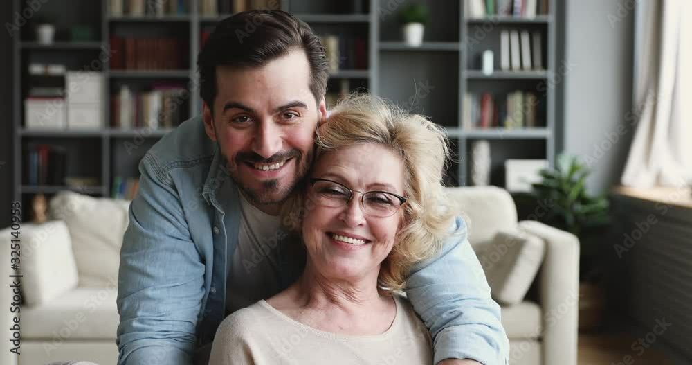 Smiling 30s son looking at camera hugging older mom indoors