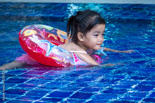 Portrait of cute little asian girl having fun in swimming pool, floating in blue refreshing water with colurful rubber ring