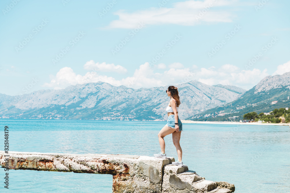 young woman traveler in short jeans shorts walking along the ocean with mountains on background young woman traveler in short jeans shorts walking along the ocean with mountains on background