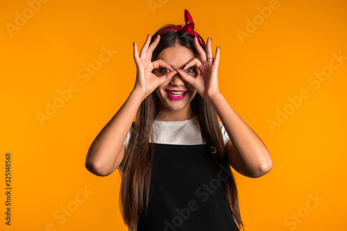 Positive girl making OK sign over eyes like glasses on yellow background and smiles to camera. Body language. Young woman.