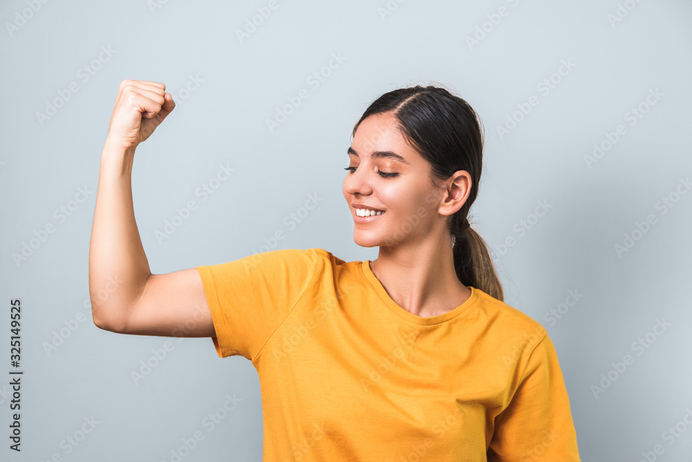 women are strong. young attractive brunette in yellow t-shirt showing her biceps on grey background women are strong. young attractive brunette in yellow t-shirt showing her biceps on grey background