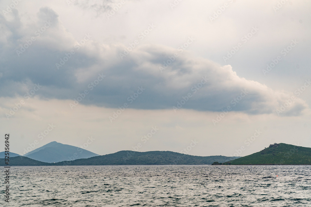 In front grey sea with little waves. Background with green hilly island and a huge cloud on grey sky In front grey sea with little waves. Background with green hilly island and a huge cloud on grey sky