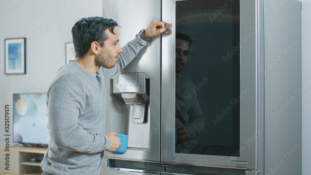 Handsome Young Man is Standing Next to a Refrigerator While Drinking His Morning Coffee. He is Checking a To Do List on a Smart Fridge at Home. Kitchen is Bright and Cozy. Handsome Young Man is Standing Next to a Refrigerator While Drinking His Morning Coffee. He is Checking a To Do List on a Smart Fridge at Home. Kitchen is Bright and Cozy.