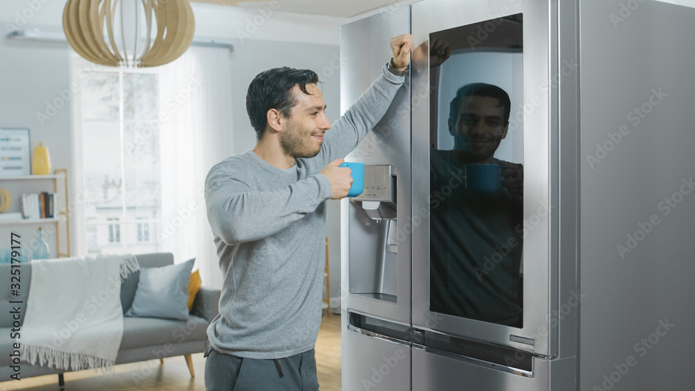 Handsome Young Man is Standing Next to a Refrigerator While Drinking His Morning Coffee. He is Checking a To Do List on a Smart Fridge at Home. Kitchen is Bright and Cozy. Handsome Young Man is Standing Next to a Refrigerator While Drinking His Morning Coffee. He is Checking a To Do List on a Smart Fridge at Home. Kitchen is Bright and Cozy.