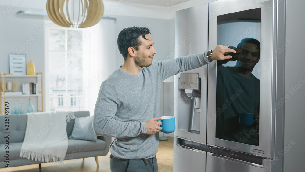 Handsome Young Man is Standing Next to a Refrigerator While Drinking His Morning Coffee. He is Checking a To Do List on a Smart Fridge at Home. Kitchen is Bright and Cozy.