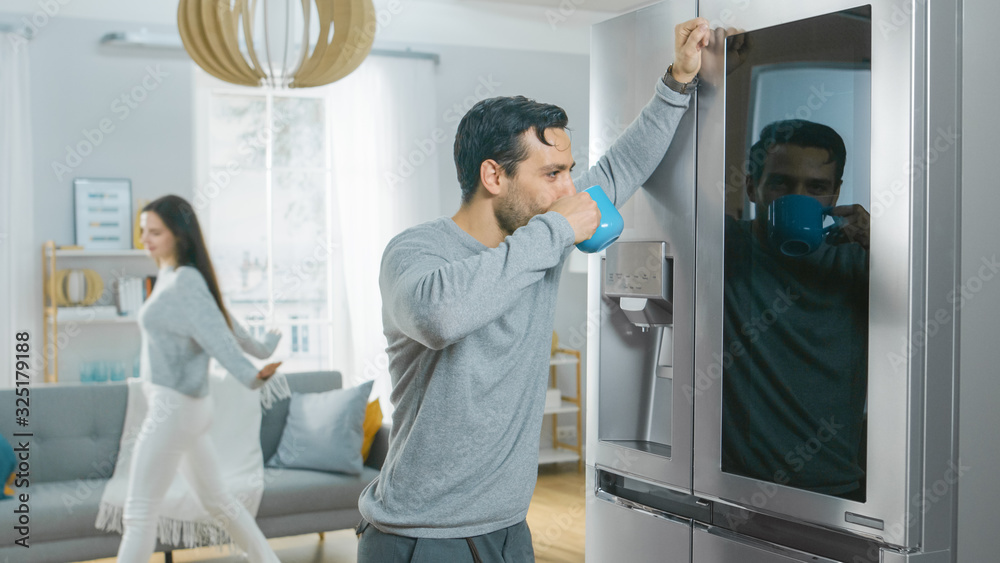 Handsome Young Man is Standing Next to a Refrigerator While Drinking His Morning Coffee. He is Checking a To Do List on a Smart Fridge at Home. Kitchen is Bright and Cozy. Handsome Young Man is Standing Next to a Refrigerator While Drinking His Morning Coffee. He is Checking a To Do List on a Smart Fridge at Home. Kitchen is Bright and Cozy.