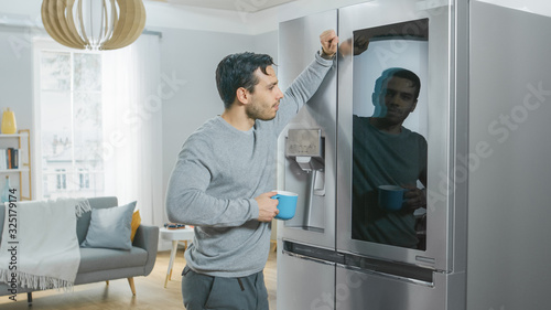 Handsome Young Man is Standing Next to a Refrigerator While Drinking His Morning Coffee. He is Checking a To Do List on a Smart Fridge at Home. Kitchen is Bright and Cozy.