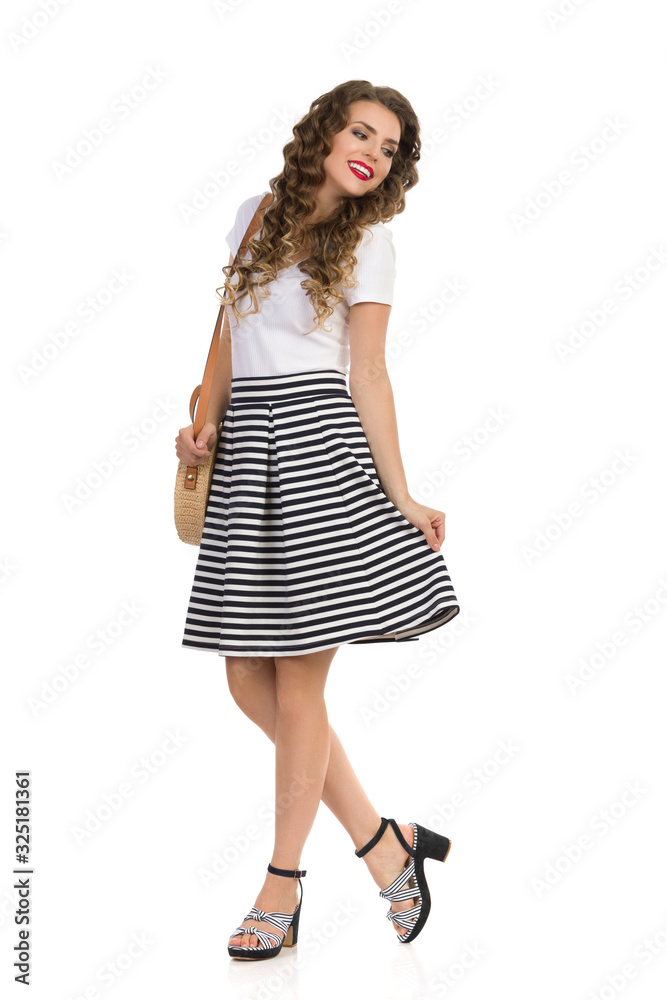 Young Woman In Striped Skirt And High Heels Is Posing With Straw Purse And Looking Away. Young Woman In Striped Skirt And High Heels Is Posing With Straw Purse And Looking Away.