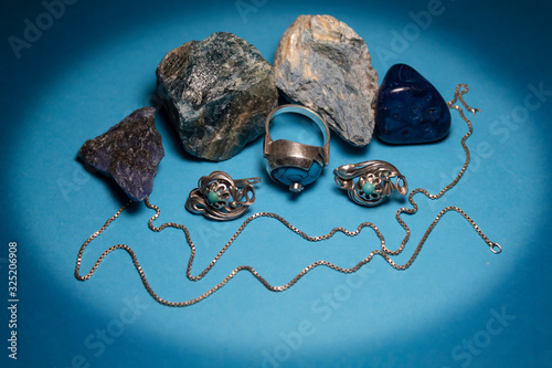 On a blue background in an oval frame are semiprecious stones agate, dumortierite, apatite, saddle and silver earrings, a ring and a chain. photo