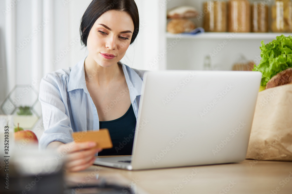 Smiling woman online shopping using computer and credit card in kitchen Smiling woman online shopping using computer and credit card in kitchen