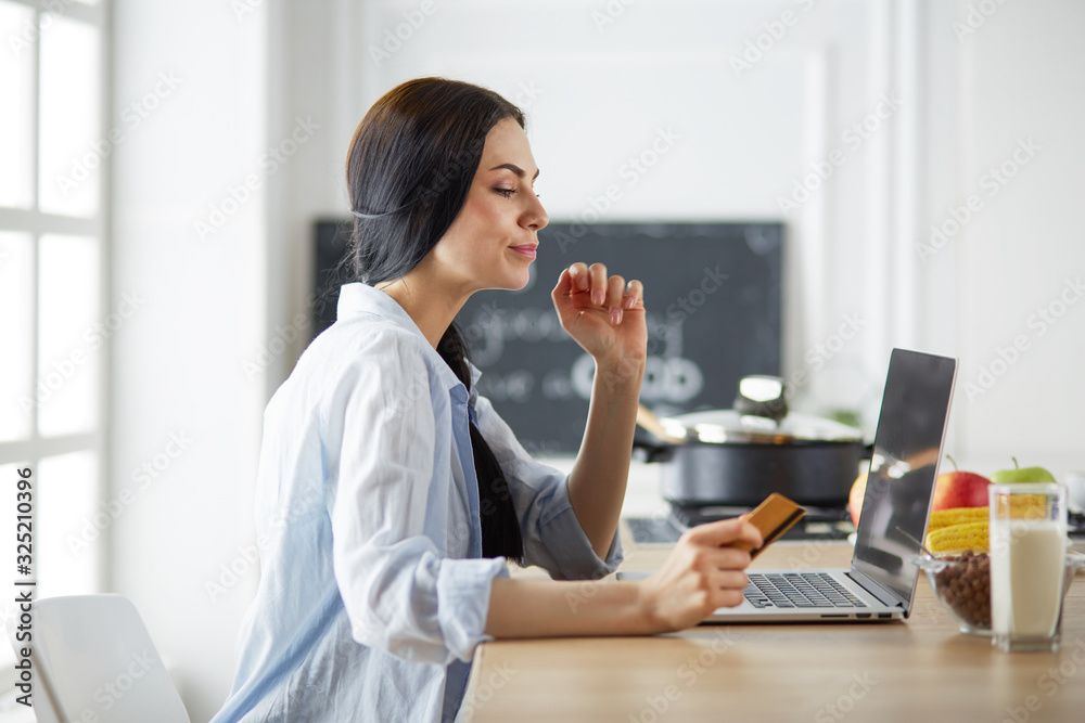 Smiling woman online shopping using computer and credit card in kitchen Smiling woman online shopping using computer and credit card in kitchen