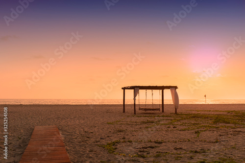 Wooden walkway to the sand beach with hammock cottage decorated with white fabric over sea water and colorful sunset sky background in Baja California, Mexico.