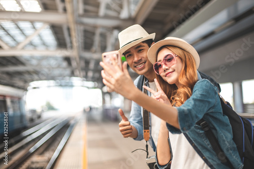 Happy Asian couple traveler holding a mobile phone in station and waiting for train in vacation time.Two Asian Tourists With Backpacks Train travel in Sightseeing City Thailand.