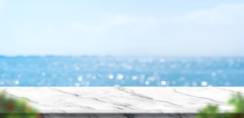Empty white marble table with blur blue sky and sea bokeh background with green leaf foreground,Mock up template for display or montage of product or content use as banner in social media ads