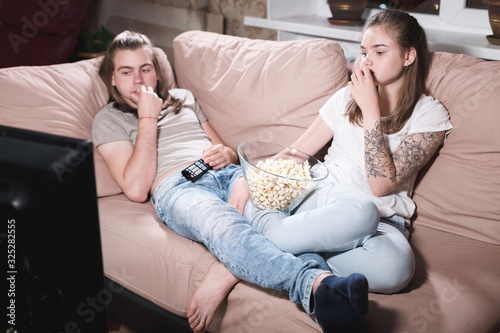 Portrait of a long-haired guy and girl with popcorn in their hands on the couch watching TV. Family evening of a young couple at the televisor. Generation of the Mellineals © yanik88