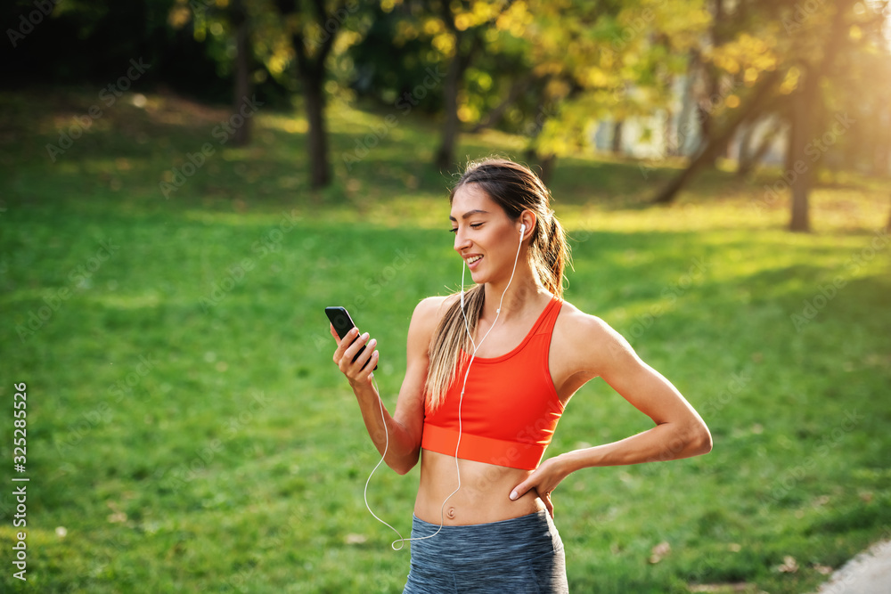 Young charming smiling sportswoman standing in public park, taking a break and listening music over smart phone. Young charming smiling sportswoman standing in public park, taking a break and listening music over smart phone.