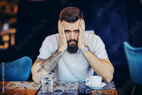 Young desperate bearded tattooed muscular handsome hipster holding hands on face and looking at camera. He is waiting for his girlfriend. photo