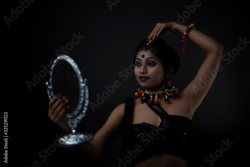 Fashion portrait of Indian Bengali brunette woman in Indian traditional tribal dress and handmade ornaments doing makeup in dark copy space studio background. Indian lifestyle and fashion photography.