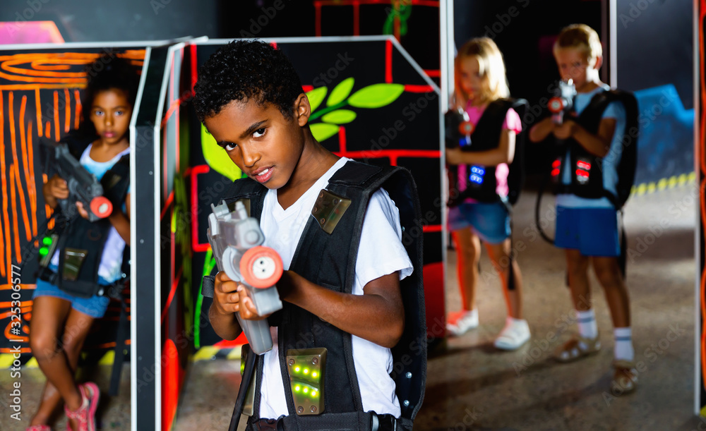 Obraz premium Emotional afro-american boy with laser pistol playing laser tag with friends on dark labyrinth