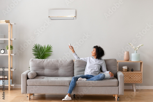 Happy african woman sitting on sofa switching on air conditioner