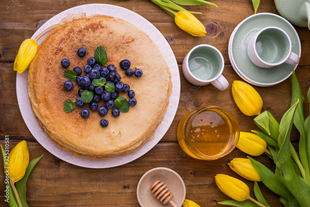 Large traditional Russian pancakes with berries and honey on a wooden table. Baking for the spring holiday Shrovetide and a bouquet of yellow tulips. Copy space Large traditional Russian pancakes with berries and honey on a wooden table. Baking for the spring holiday Shrovetide and a bouquet of yellow tulips. Copy space