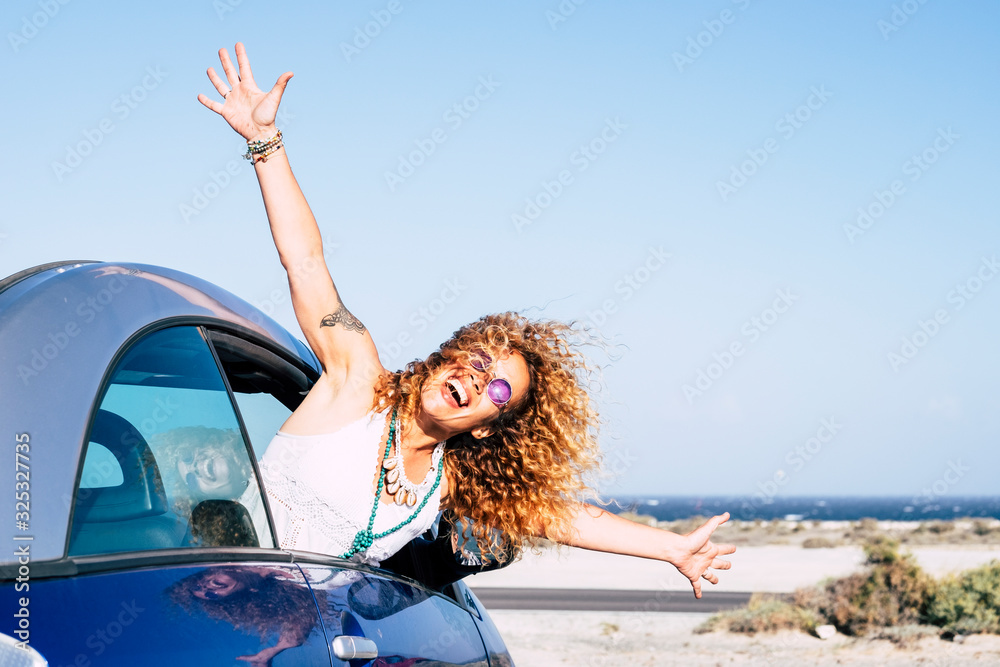 Happy and joyful people in outdoor travel leisure activity - beautiful curly blonde woman have fun outside the window of a blue vehicle car - concept of transport and happiness - ocean background - obrazy, fototapety, plakaty 