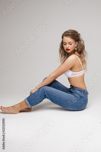 Attractive young barefoot lady in white bra and jeans sitting on floor isolated on grey background while looking at camera