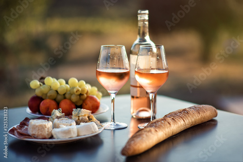 Bottle of rose wine and two full glasses of wine on table in heart of Provence, France with french bread, cheese, ham, grapes and peaches with olive trees on background in sunset. Travel in Provence.