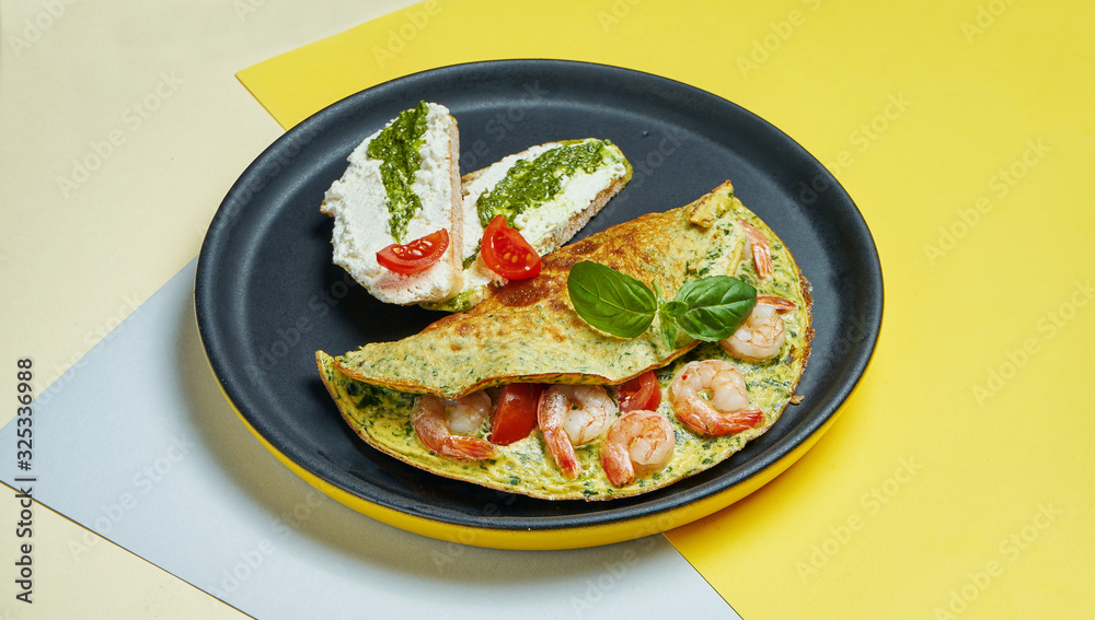 Continental breakfast - omelet with shrimps, tomatoes and basil with toast with cream cheese and pesto on a black plate on colored backgrounds.