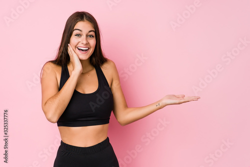 Fototapeta Naklejka Na Ścianę i Meble -  Young caucasian fitness woman doing sport isolated holds copy space on a palm, keep hand over cheek. Amazed and delighted.