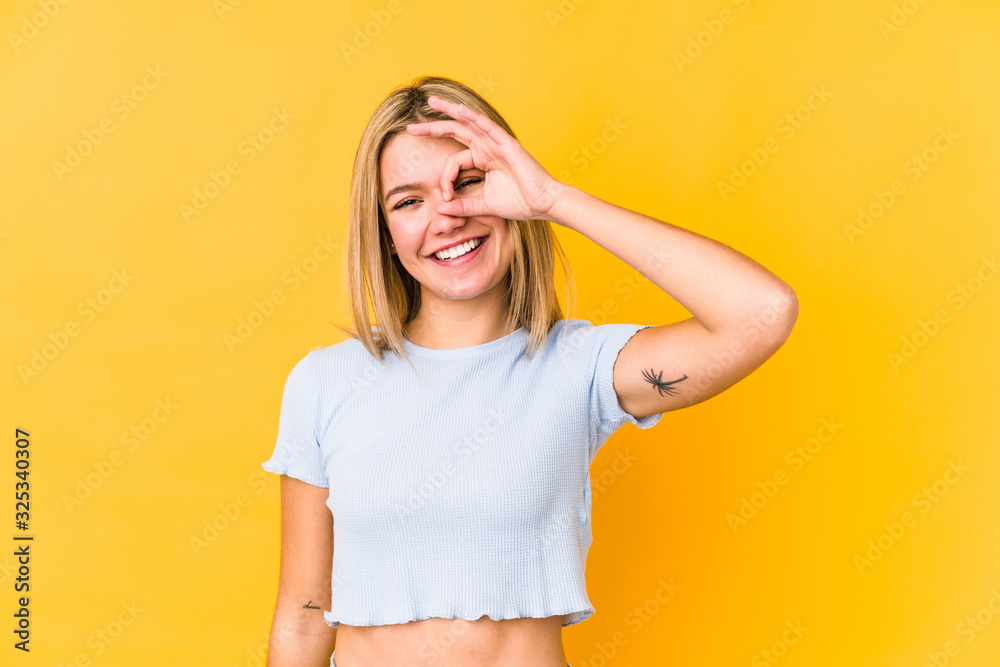 Young blonde caucasian woman isolated on a yellow background excited keeping ok gesture on eye. Young blonde caucasian woman isolated on a yellow background excited keeping ok gesture on eye.