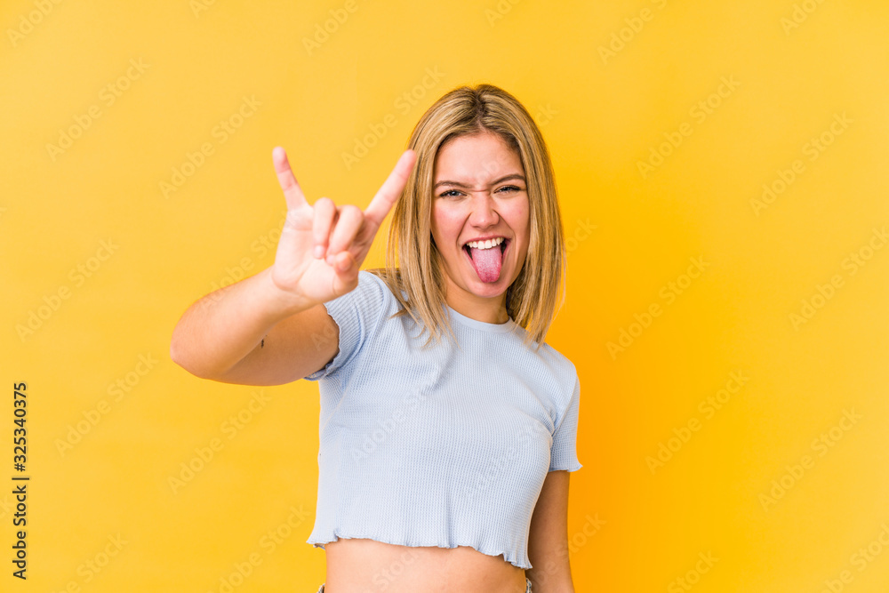 Young blonde caucasian woman isolated on a yellow background showing a horns gesture as a revolution concept.
