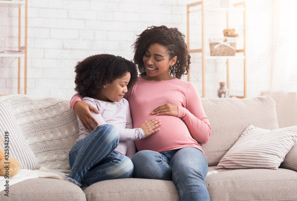 Curious little daughter touching her pregnant mother tummy, sitting on sofa at home, empty space