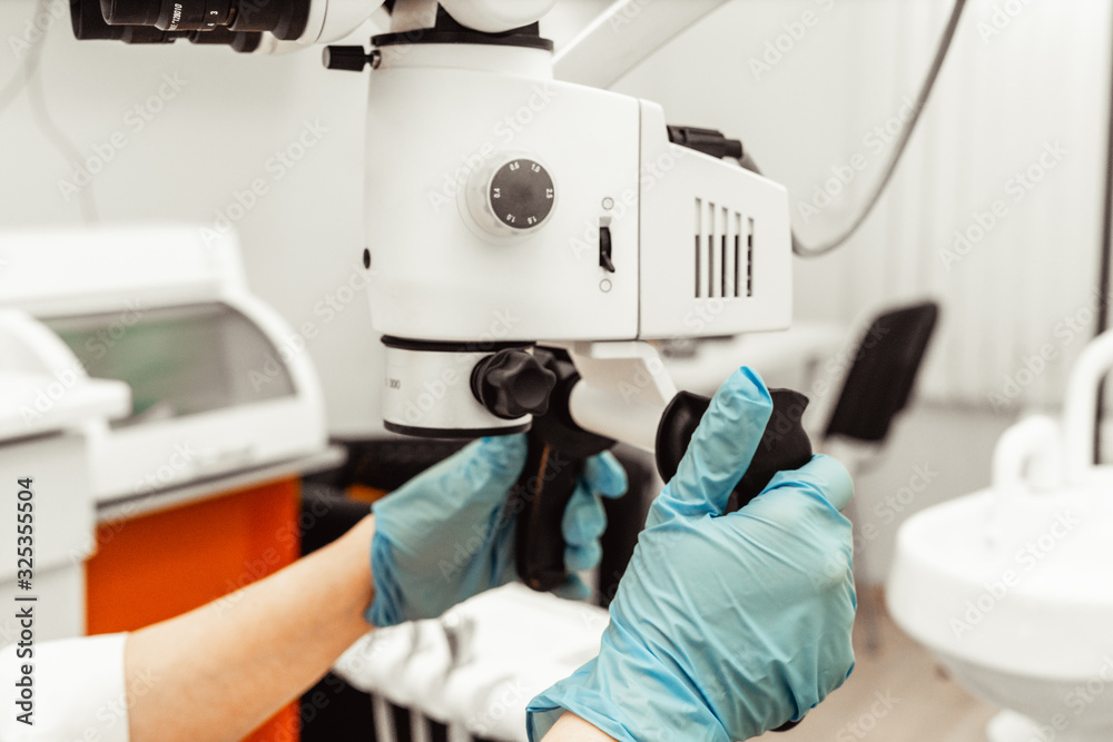 Young woman dentist doctor looks through a professional microscope in a dental clinic. A doctor in a disposable medical mask and cap. Advanced equipment in dentistry