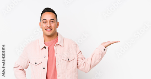 Young latin man posing isolated showing a copy space on a palm and holding another hand on waist.