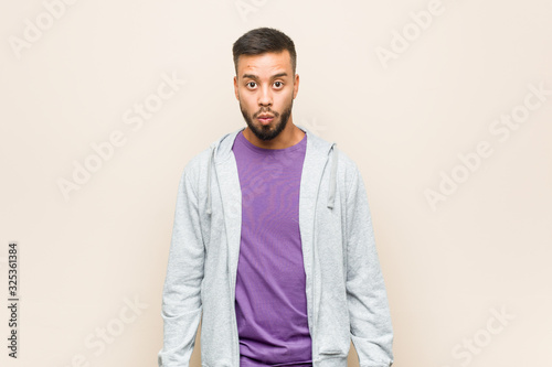 Young south-asian man shrugs shoulders and open eyes confused.