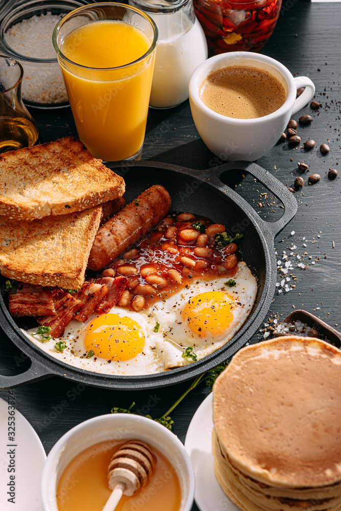 English breakfast on a dark table. fried eggs with bacon canned beans and sausage. near coffee, orange juice and pancakes with butter and honey