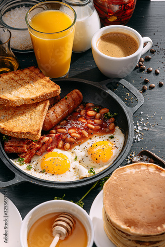 Obraz na płótnie English breakfast on a dark table. fried eggs with bacon canned beans and sausage. near coffee, orange juice and pancakes with butter and honey w salonie